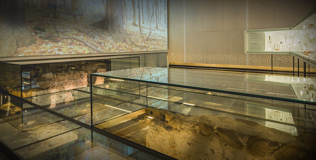 Ici a été fondée Montréal, Pointe-à-Callières, Montréal : grande image de sous-bois rétroéclairée sur le mur, vestiges de fondations sous le plancher de verre et vitrine d’objets archéologiques au fond.