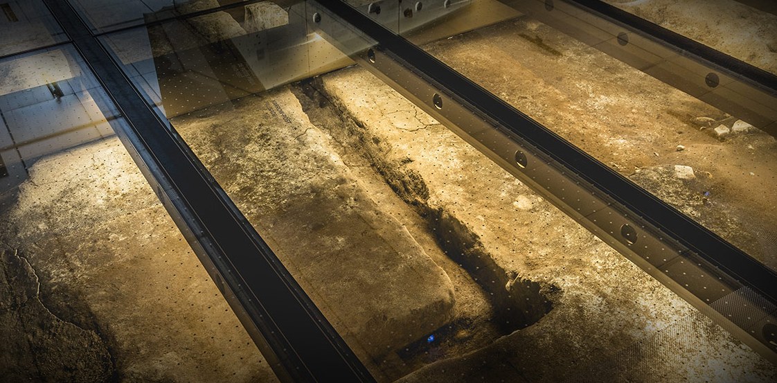Ici a été fondée Montréal, Pointe-à-Callières, Montréal : vue rapprochée du plancher de verre, la lumière rasante révèle le relief des fondations de pierre.